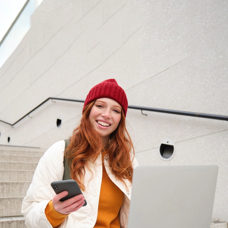 Junge Frau sitzt mit Smartphone und Laptop auf einer Treppe. 